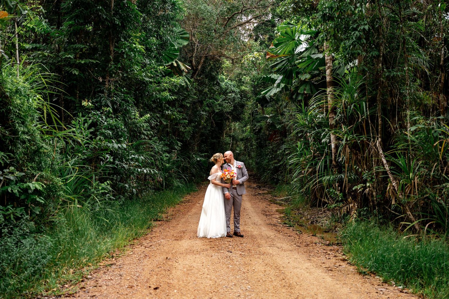 Bride and groom sharing a romantic kiss on a rustic dirt road surrounded by lush tropical rainforest.