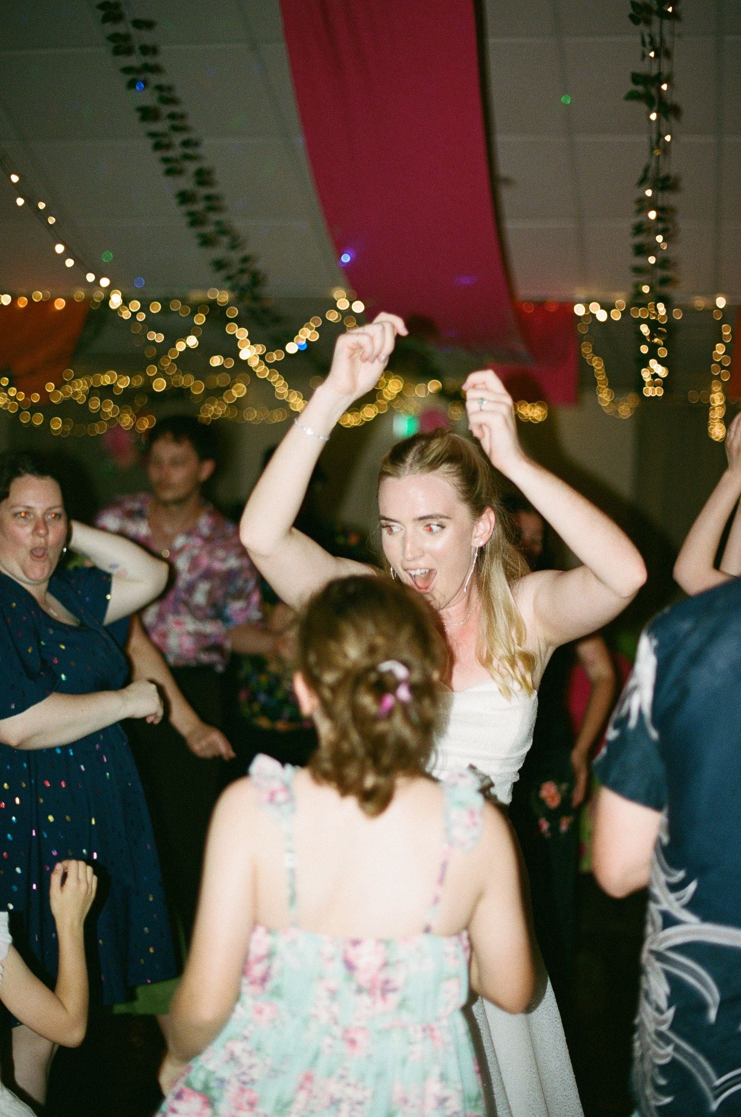 Bride in white dress dancing energetically with arms raised among guests at a wedding reception.