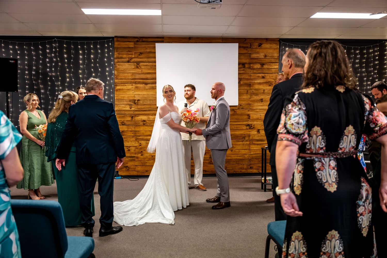 Bride and groom stand at altar during indoor wedding ceremony with wooden feature wall backdrop.