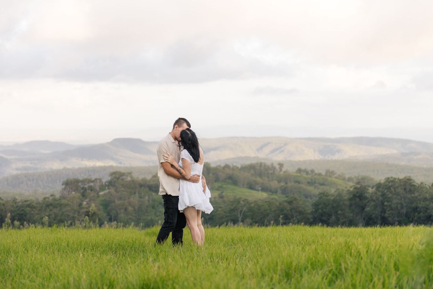 Couple sharing a tender embrace in a lush green field overlooking rolling hills.