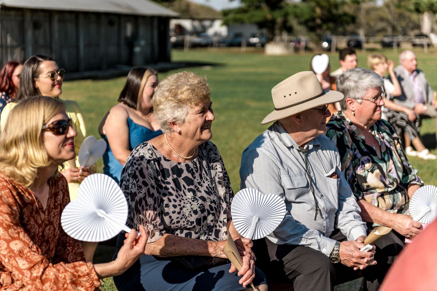 Wedding guests sitting outdoors holding white paper fans on a warm sunny day during an outdoor ceremony.