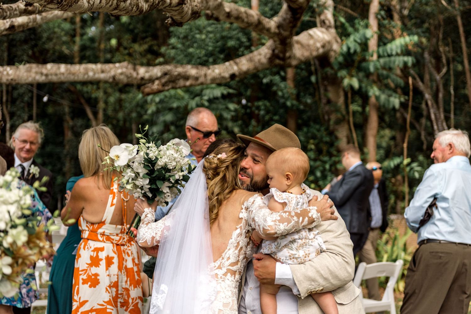 Bride hugging man holding baby while guests celebrate after outdoor wedding ceremony.