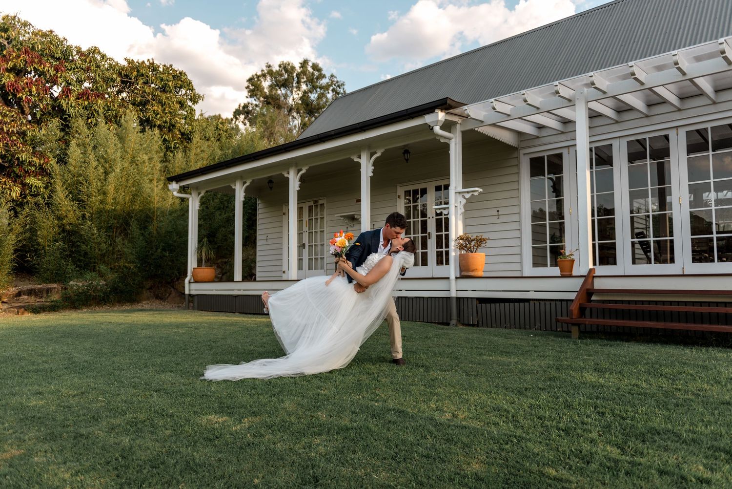 Groom dips bride dramatically on green lawn in front of a charming country homestead.