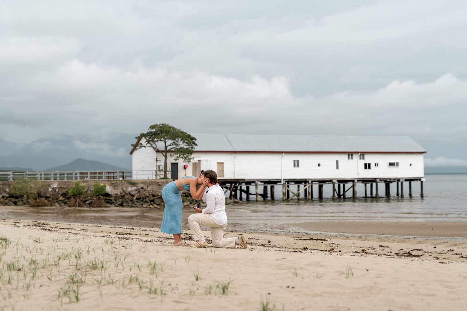 Beach marriage proposal with man on one knee before woman in blue dress near white pier.