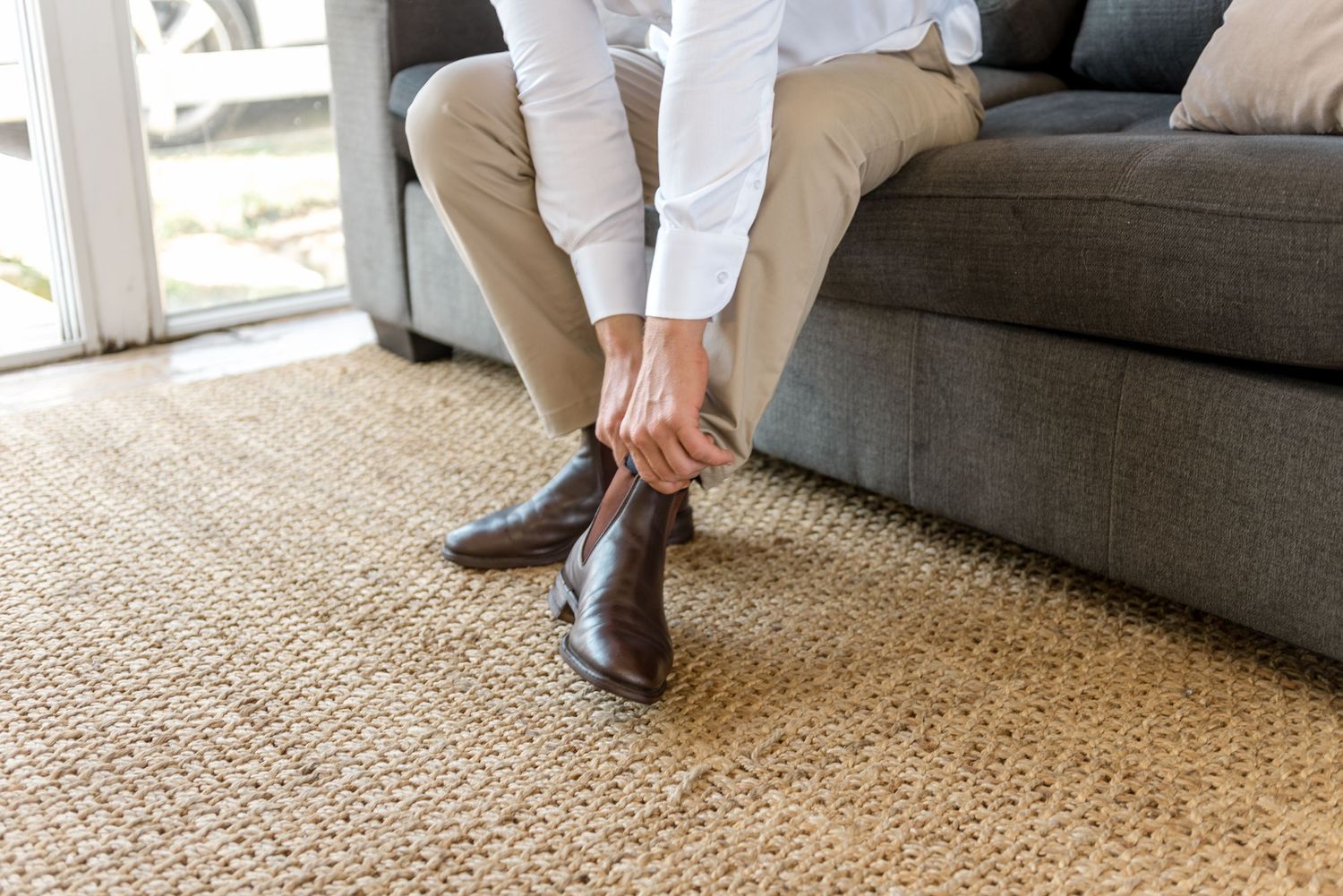 Groom putting on brown Chelsea boots while seated on grey couch during wedding morning preparations.