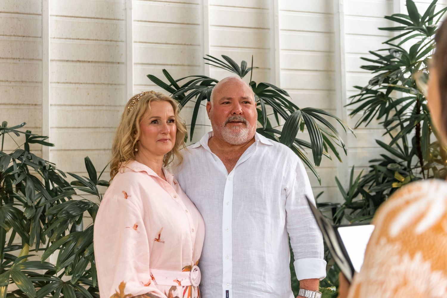 Smiling mature couple posing together in front of lush tropical plants and a white wall.