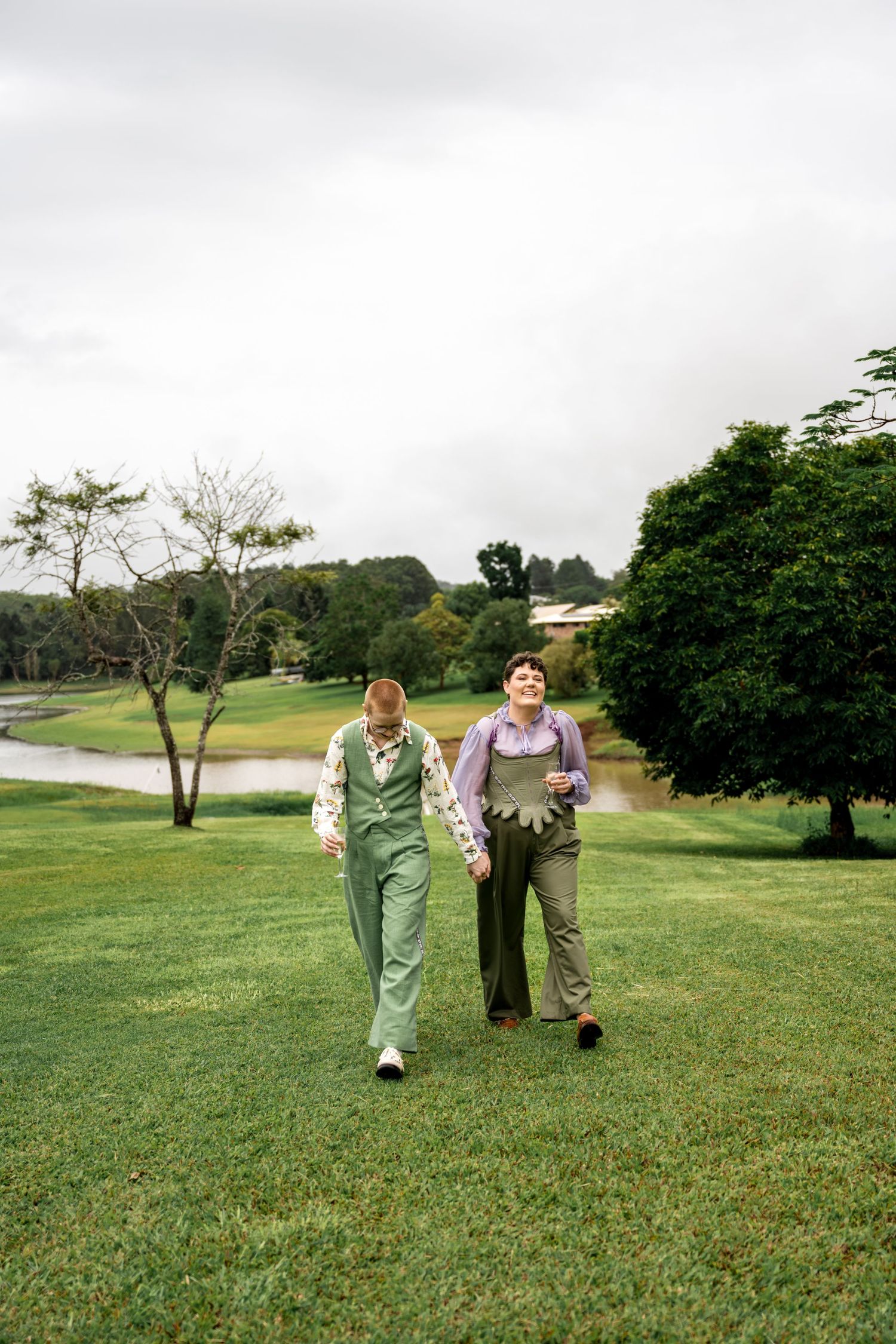 Two people in green outfits holding hands and walking across a green lawn near a pond.