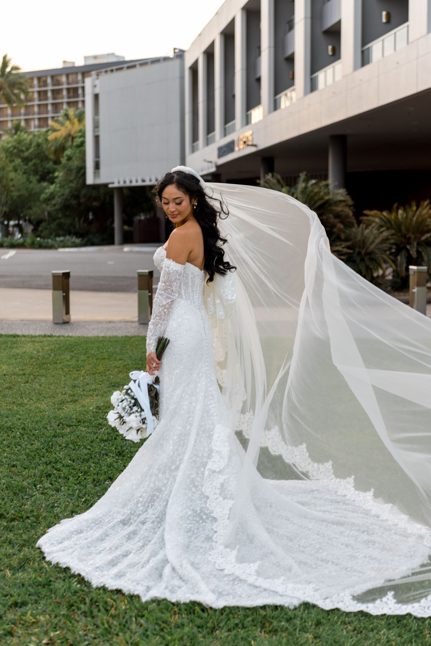 Elegant bride in fitted lace wedding dress with cathedral veil billowing in wind holding white floral bouquet outdoors.