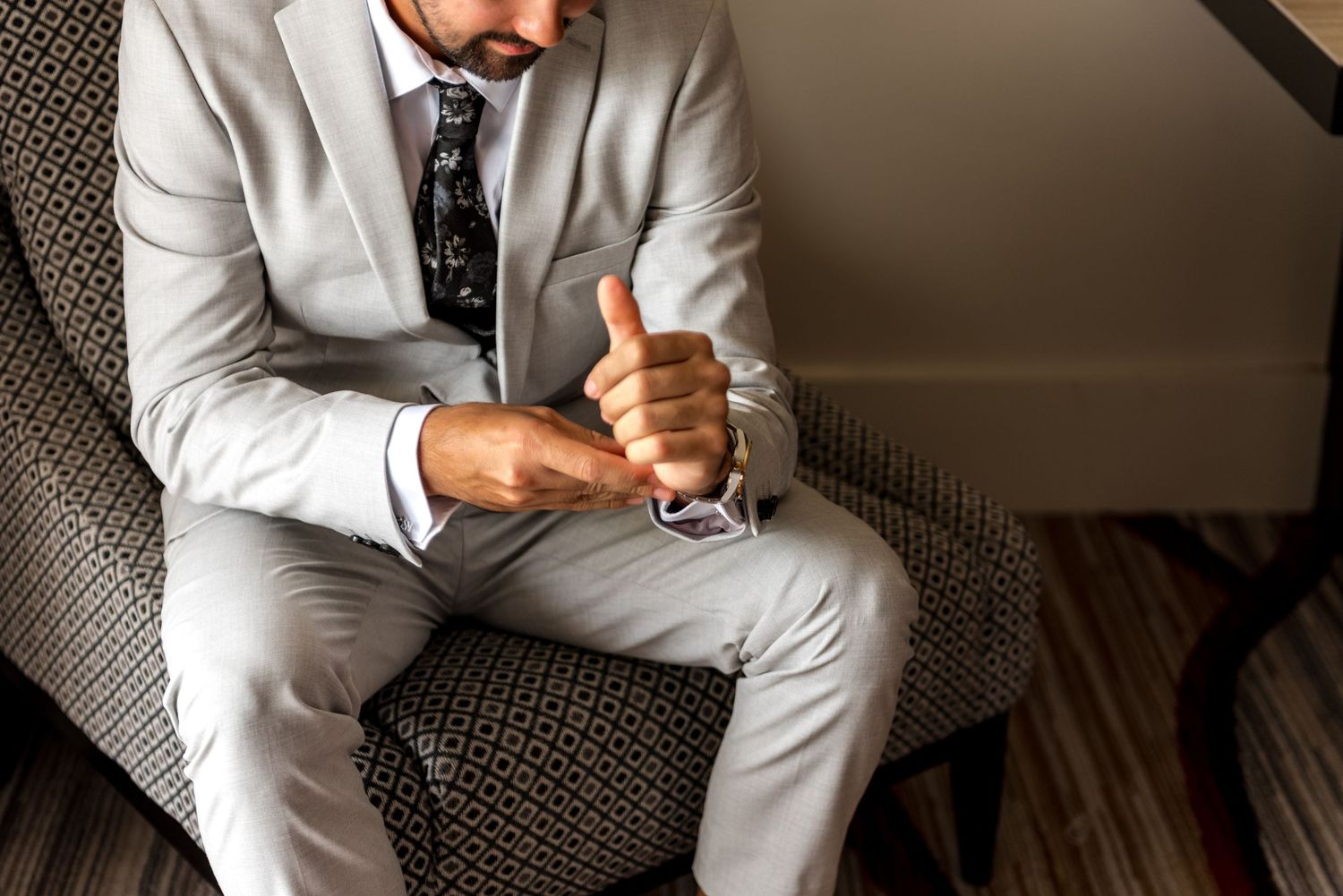 Close-up of groom in light gray suit fastening cufflinks while seated in a patterned hotel armchair.