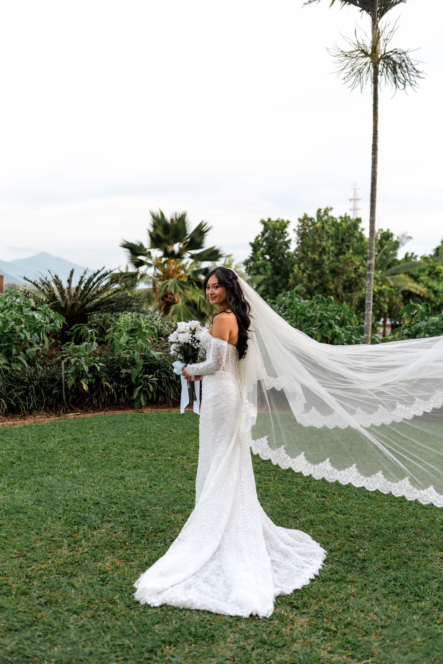 Bride in lace off-shoulder gown with flowing cathedral veil on green lawn surrounded by tropical palms.