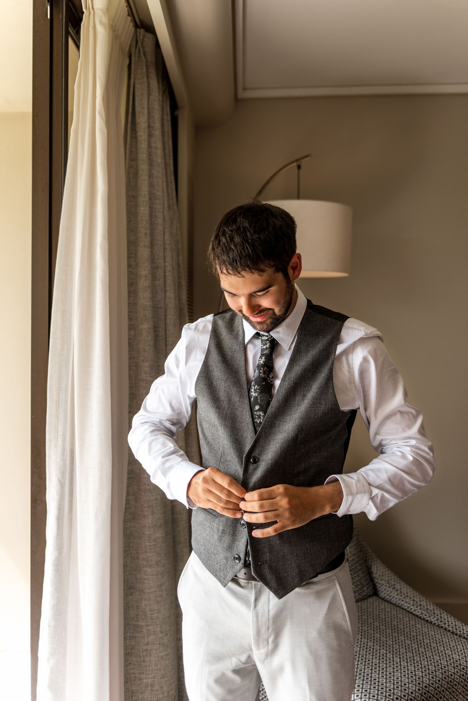 Groom in light gray suit and floral tie buttoning his vest while getting ready by a hotel window.