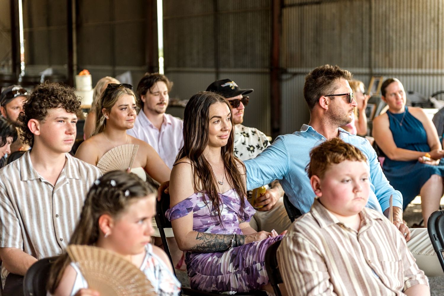Stylish wedding guests seated in barn venue, including woman in purple floral dress holding decorative fan.