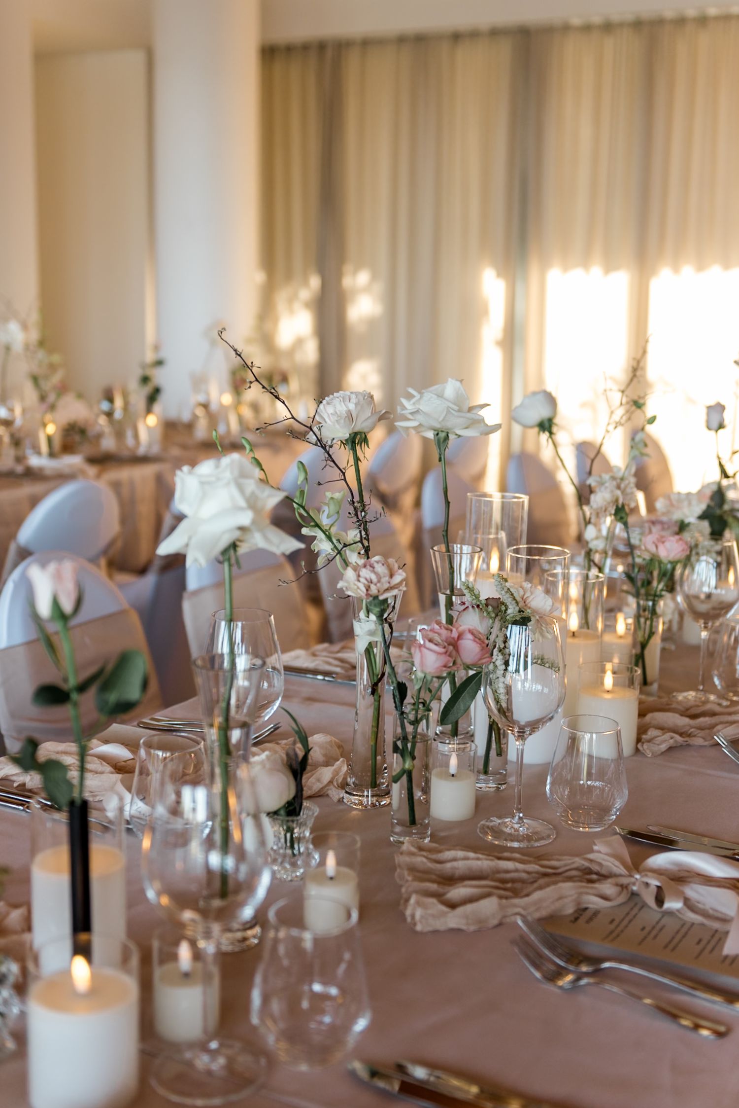 Romantic wedding reception tables adorned with white roses, pink flowers, candles, and glassware in a softly lit venue.