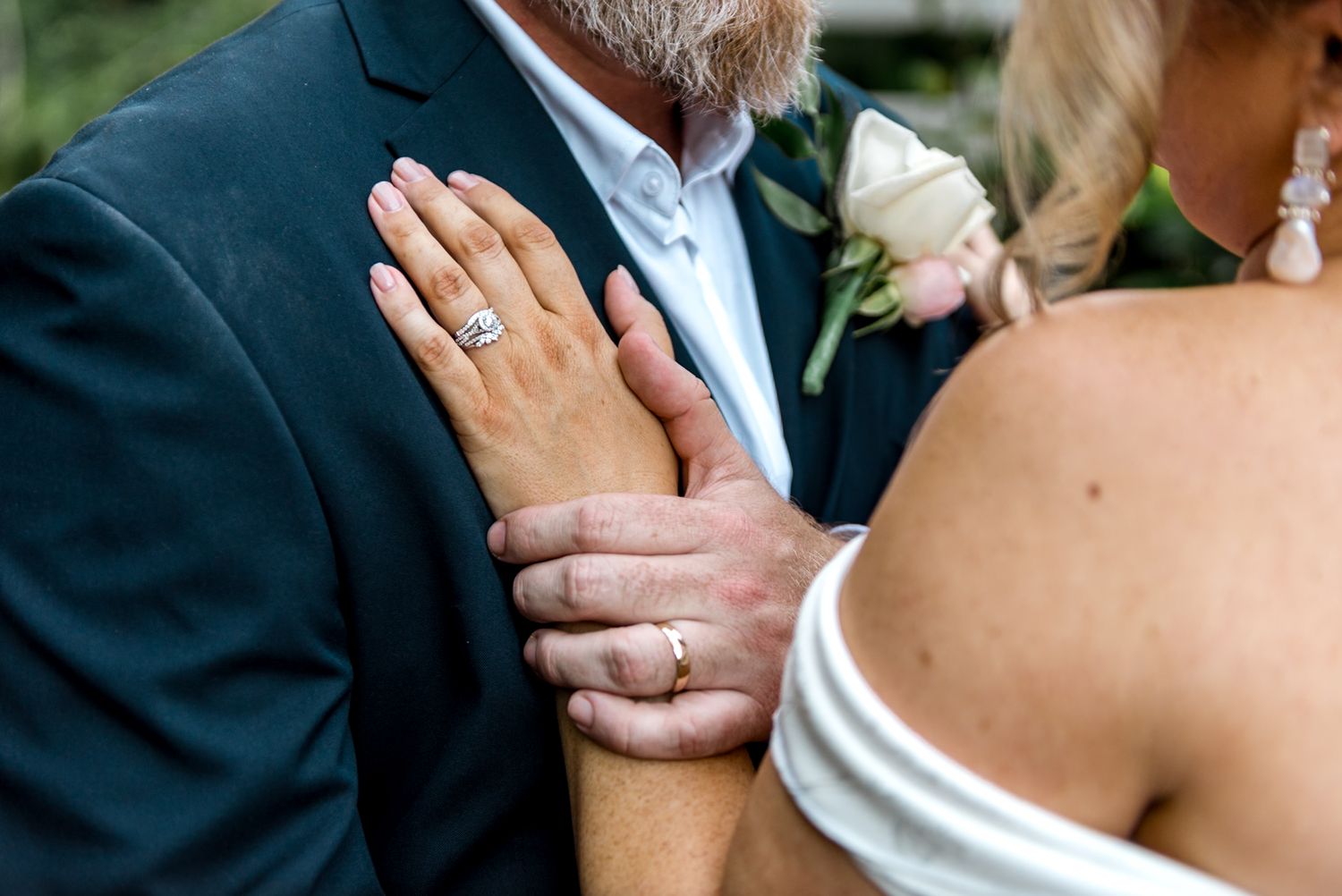 Close-up of bride and groom wedding rings as she holds his arm near boutonniere.