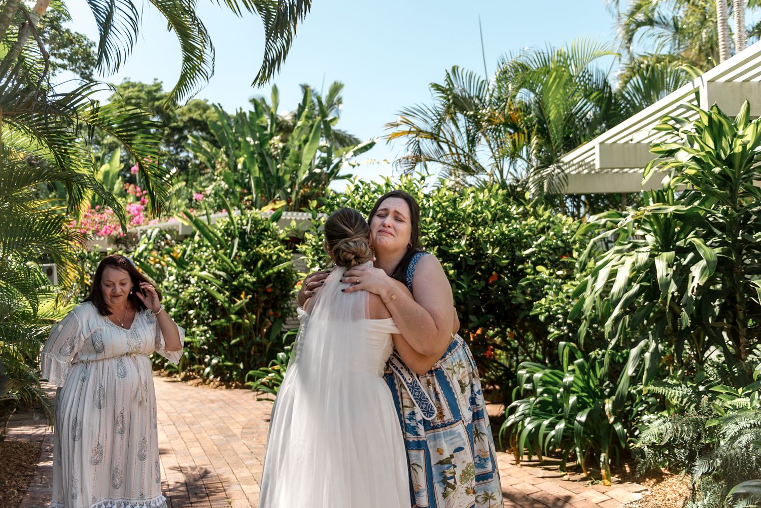 Bride in white gown embracing a guest in blue floral dress in lush tropical garden setting.