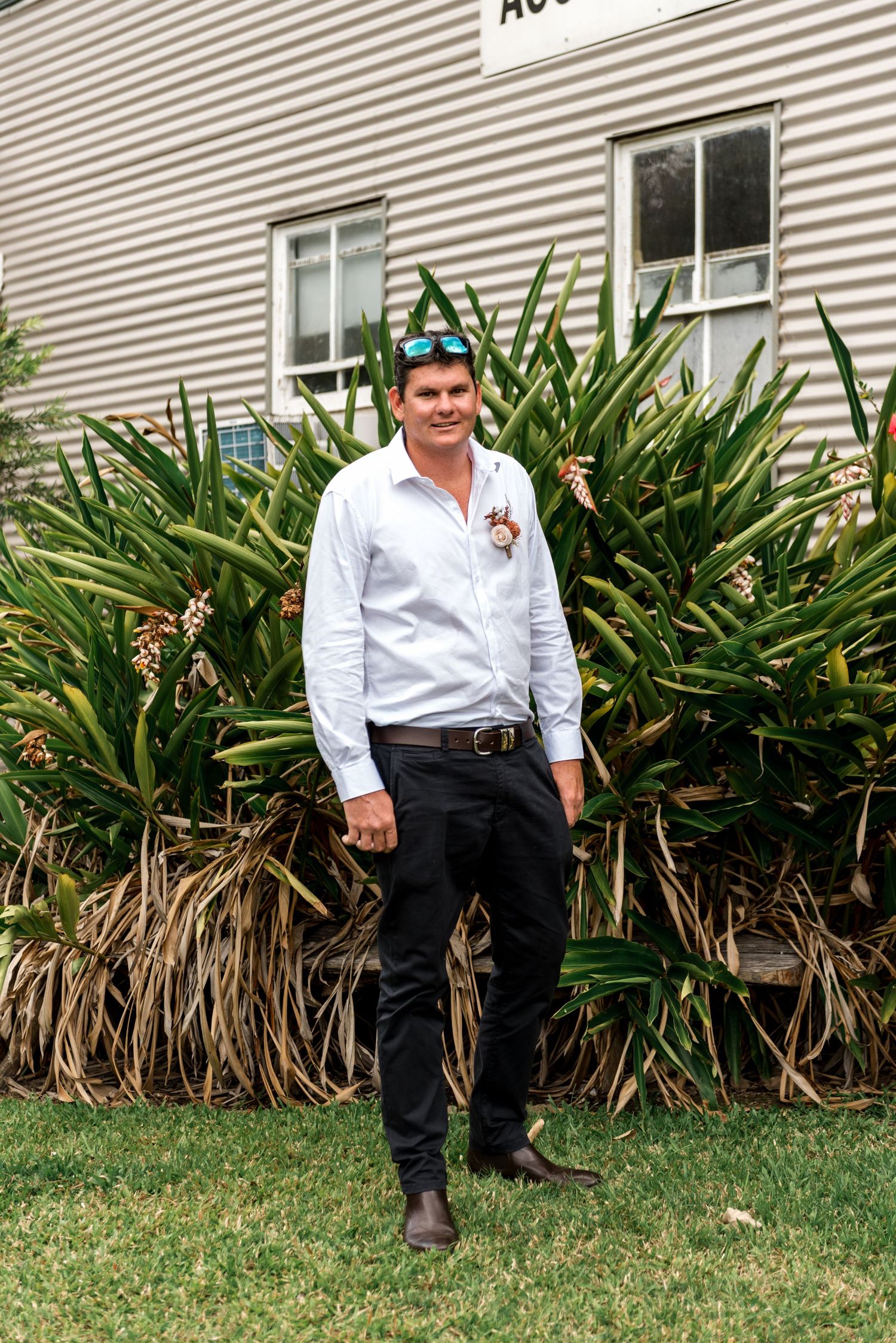 Groom in white shirt and black pants with boutonniere posing in front of tropical garden plants.