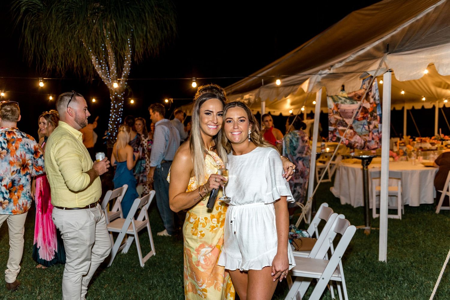 Two women smiling and posing together at an outdoor wedding reception under a white tent with string lights.