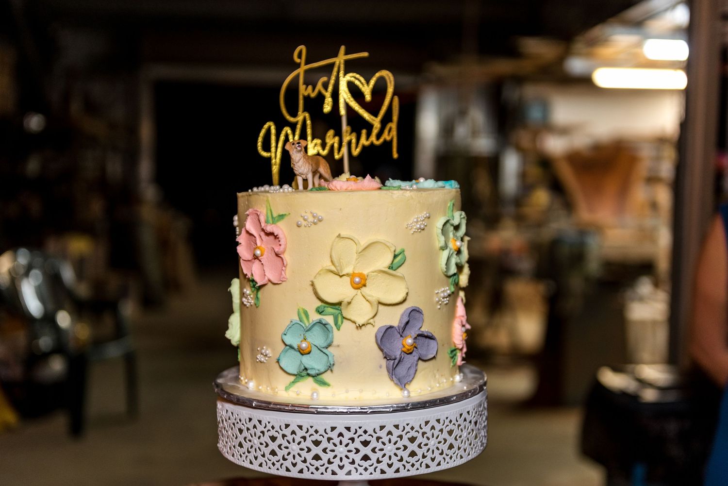 Beautiful single-tier wedding cake with colorful floral decorations and gold Just Married topper on white lace cake stand.