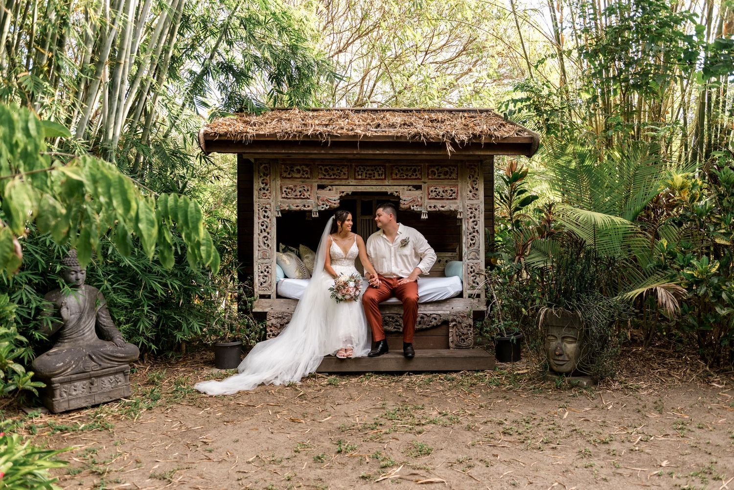 Bride and groom sit together in a rustic Balinese gazebo surrounded by tropical gardens.