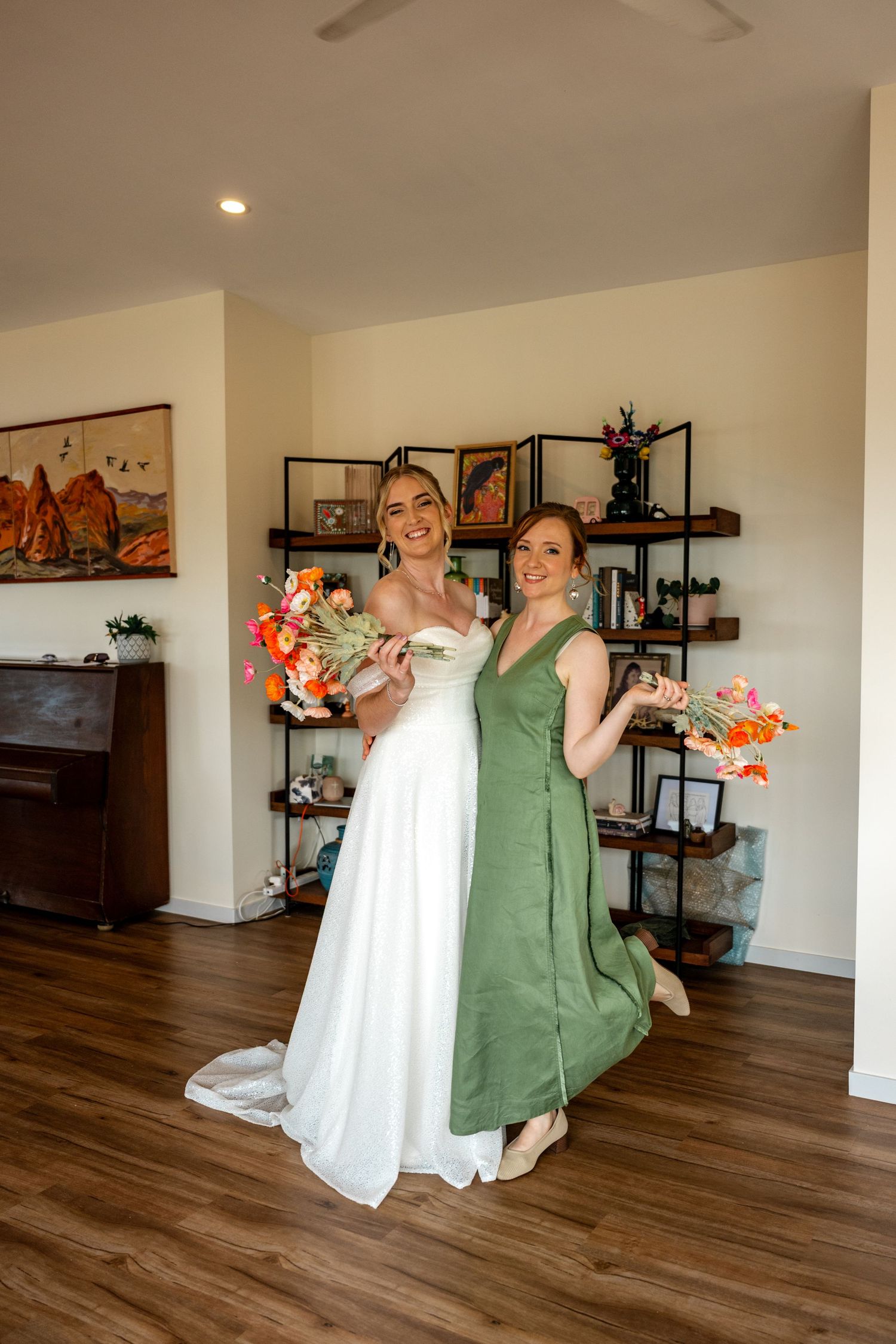 Bride in white off-shoulder gown and bridesmaid in sage green dress pose together holding vibrant orange floral bouquets.