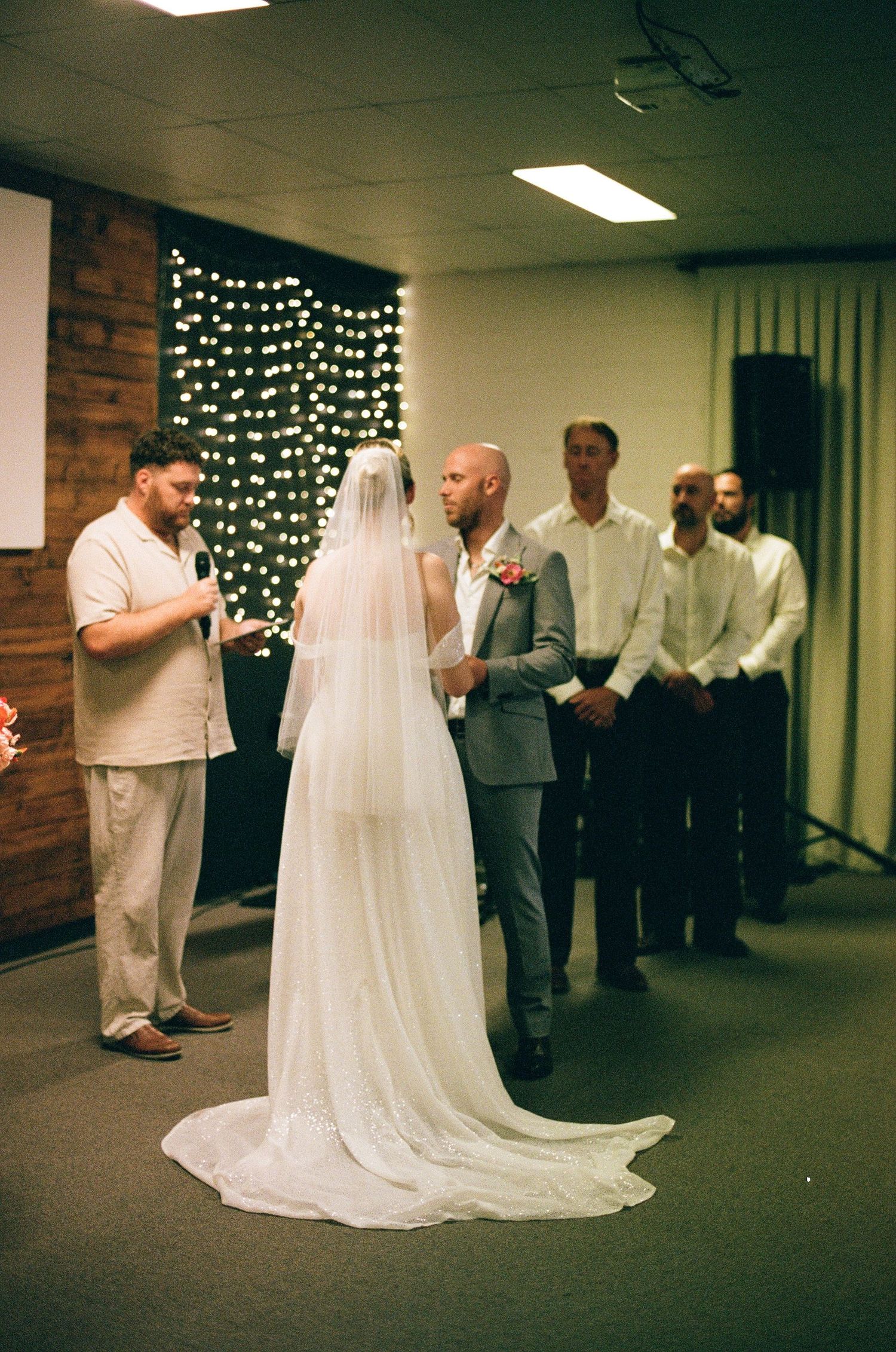 Bride and groom stand before officiant during intimate indoor wedding ceremony with fairy light backdrop and groomsmen.