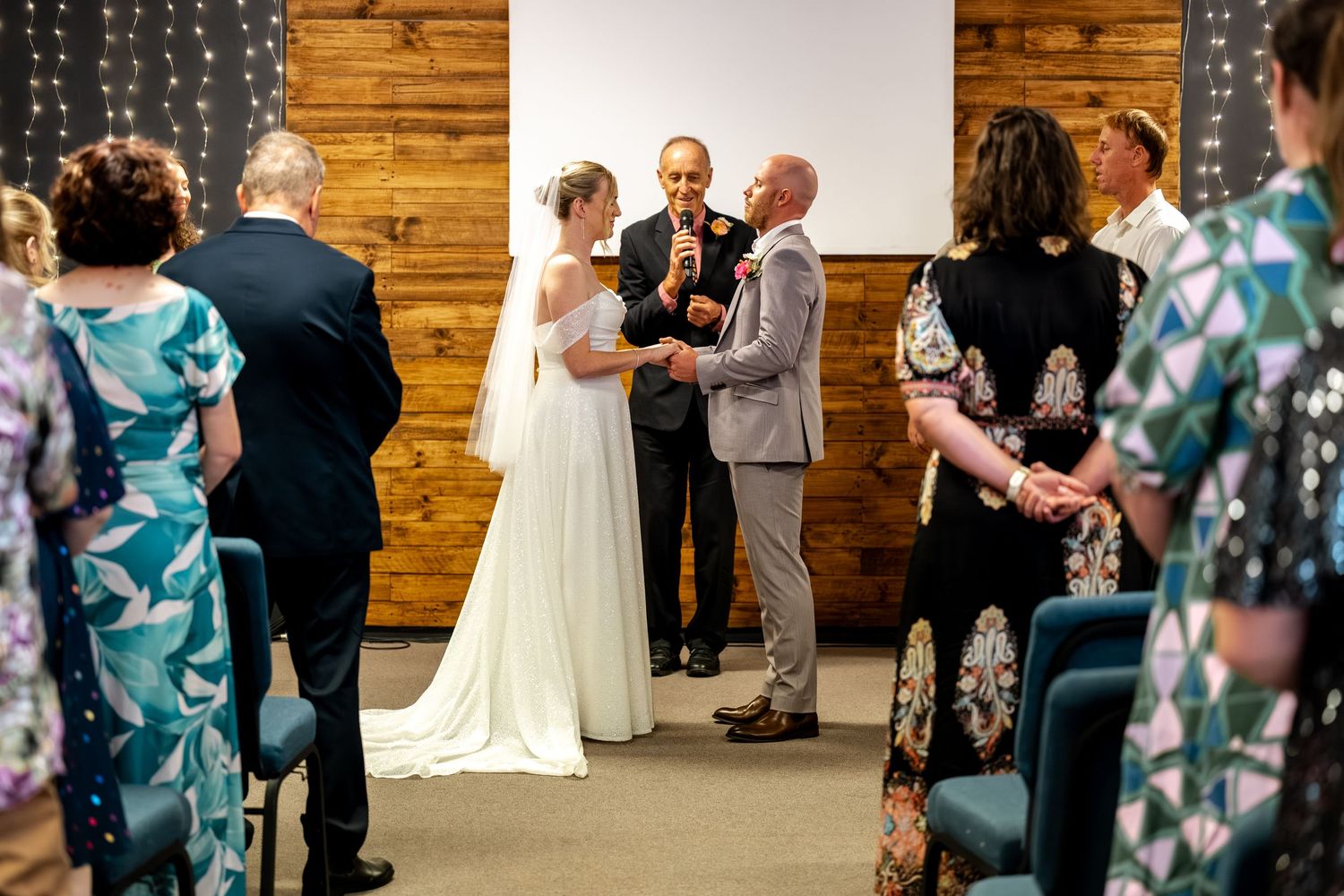 Bride and groom stand at altar during wedding ceremony with guests seated in blue chairs watching.