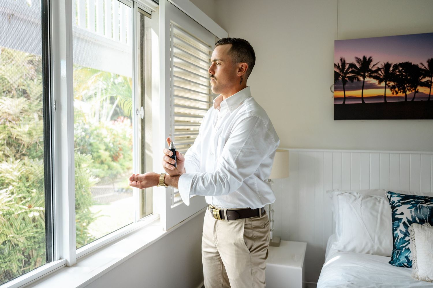 Groom in white shirt holding cologne bottle while gazing out a bright window in a bedroom.