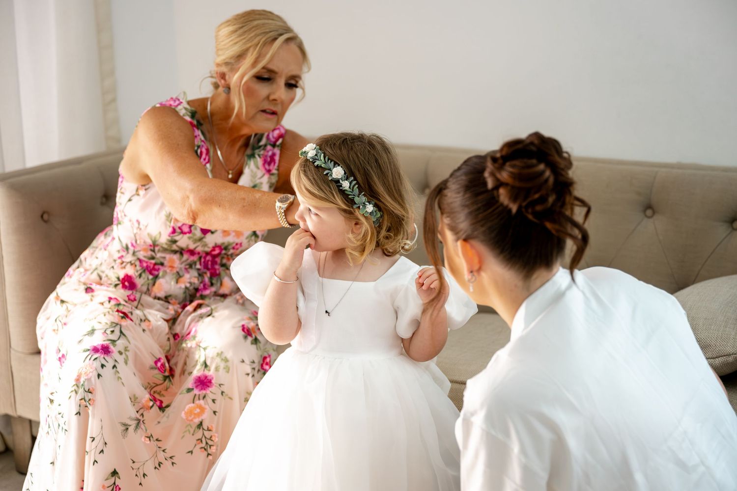 Blonde woman in floral dress helps young flower girl in white dress with floral crown at wedding preparation.