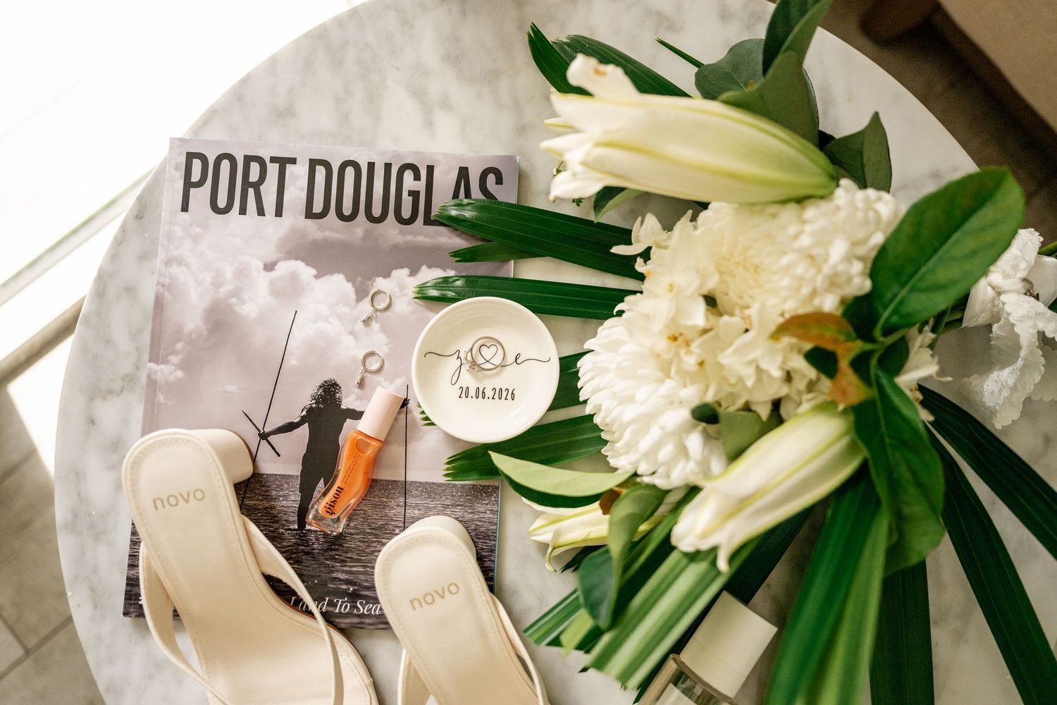 Bridal flat sandals, white lily bouquet, ring dish, and perfume on a Port Douglas marble table.