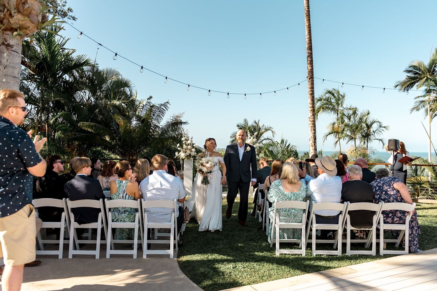 Bride walking down aisle with escort at tropical outdoor wedding ceremony with seated guests.