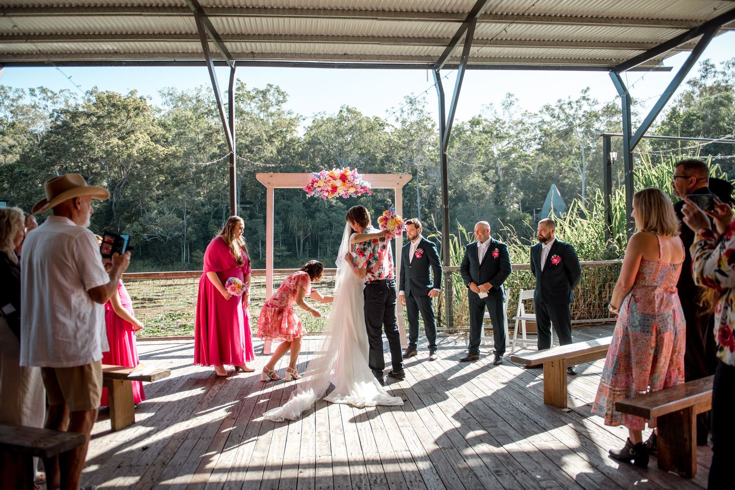 Bride walks down aisle at outdoor wedding ceremony on wooden deck surrounded by guests.