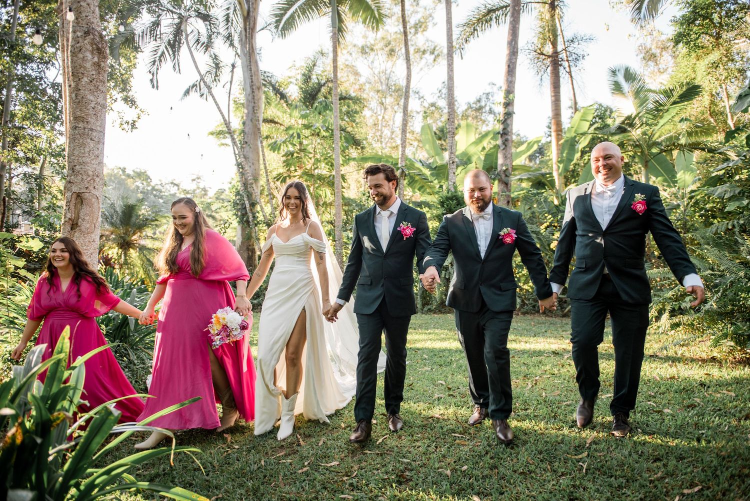 Wedding party walks together through tropical garden with bride, groom, groomsmen, and bridesmaids in pink.