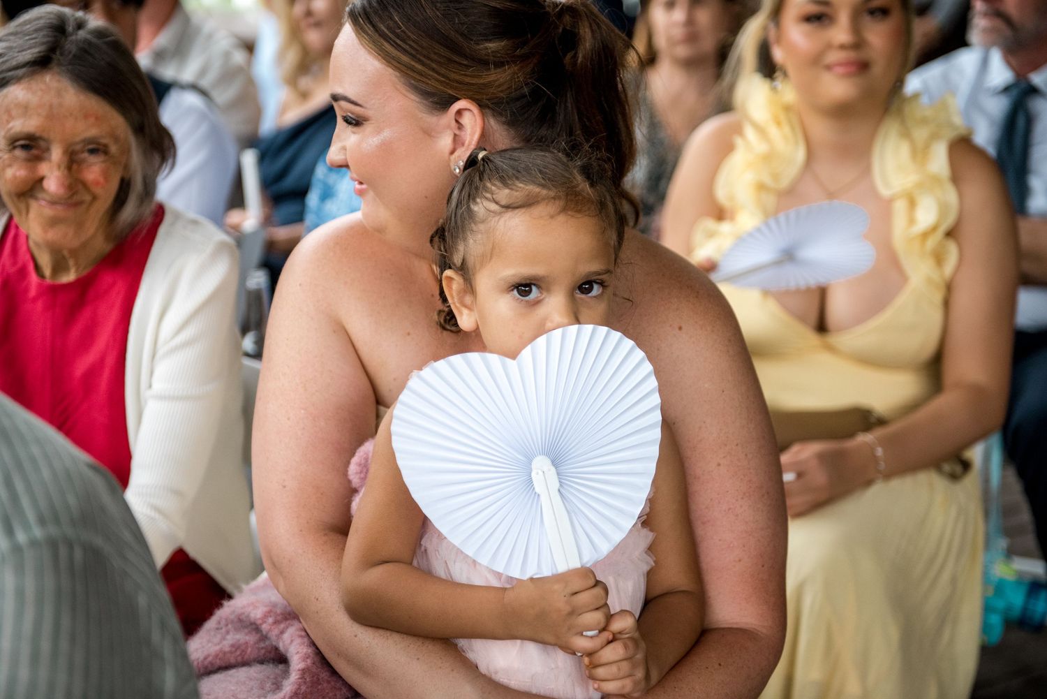 Bridesmaid in pink dress holding a young child with a white heart-shaped fan at a wedding.