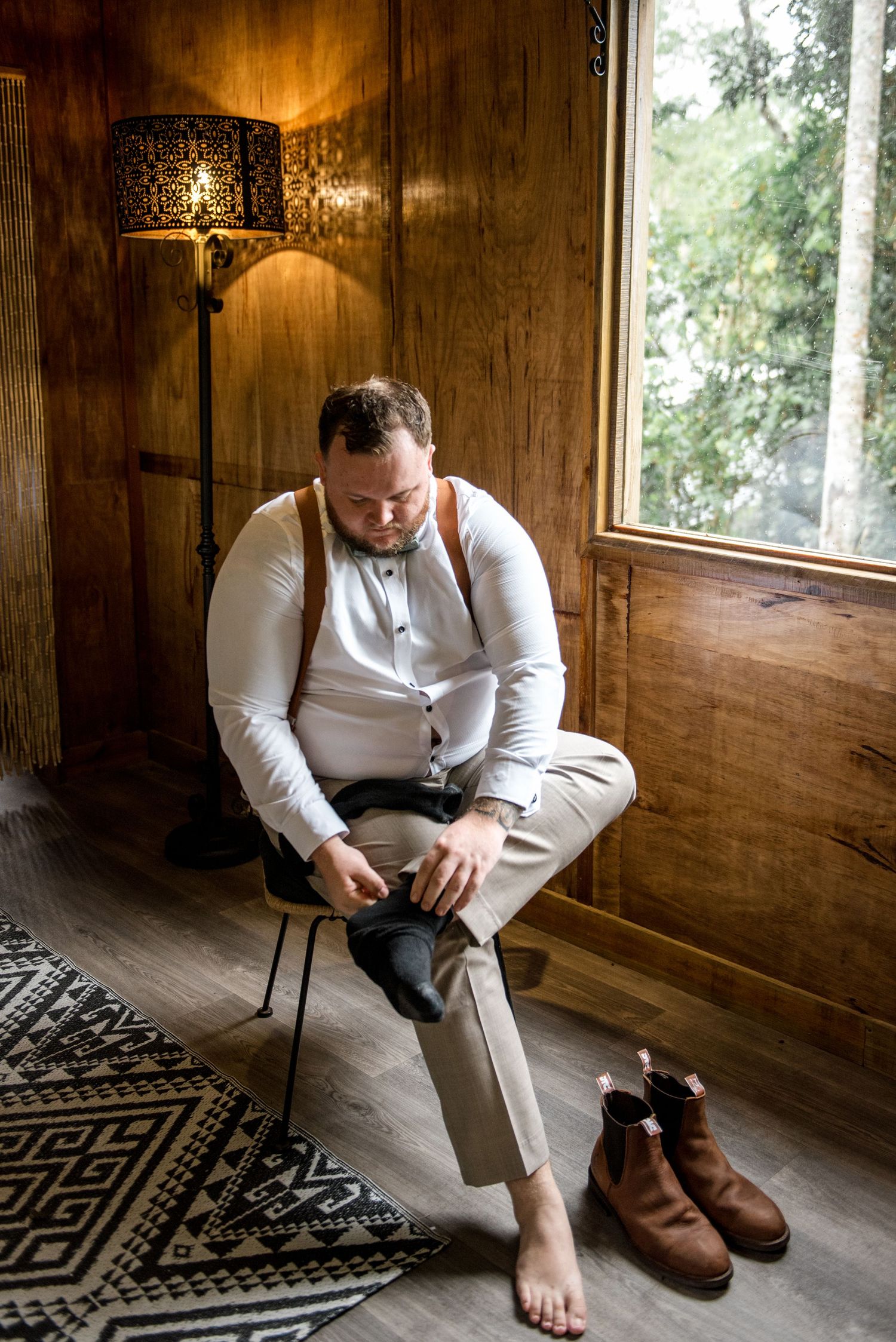 Groom in white shirt and suspenders sitting in chair putting on socks beside brown Chelsea boots.