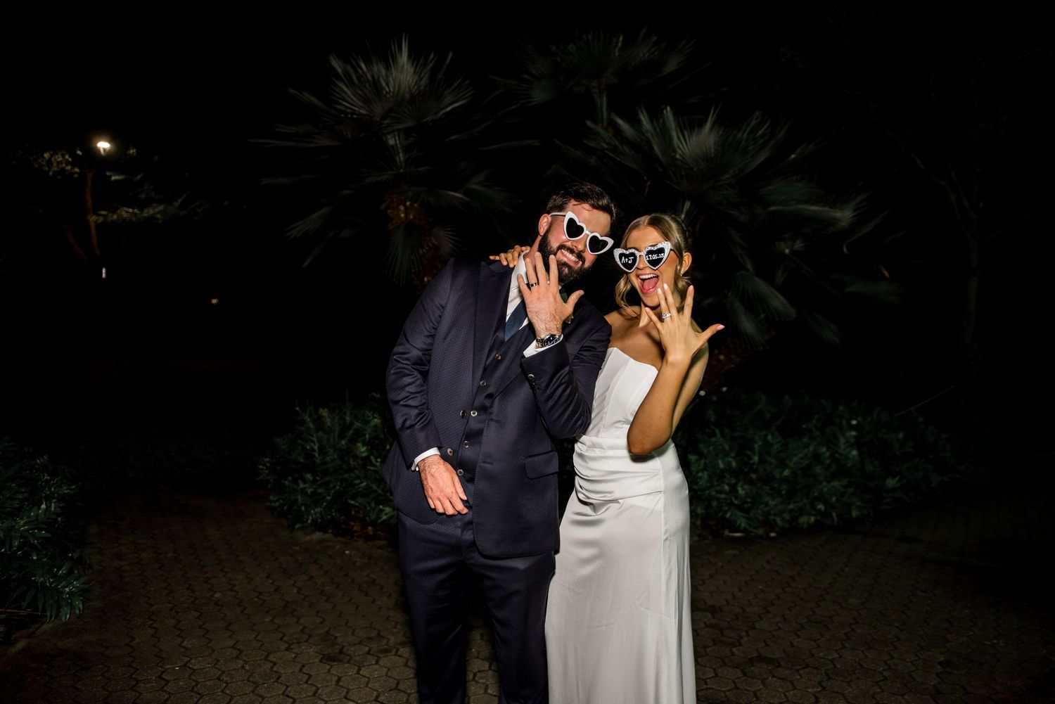 Bride and groom posing outdoors at night wearing novelty sunglasses and making fun gestures at their wedding reception.