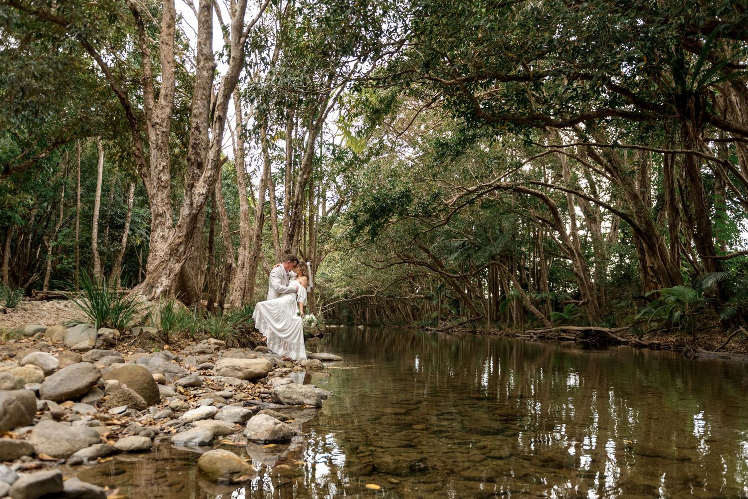 Wedding couple embrace by a tranquil forest creek with lush green trees reflected in water.