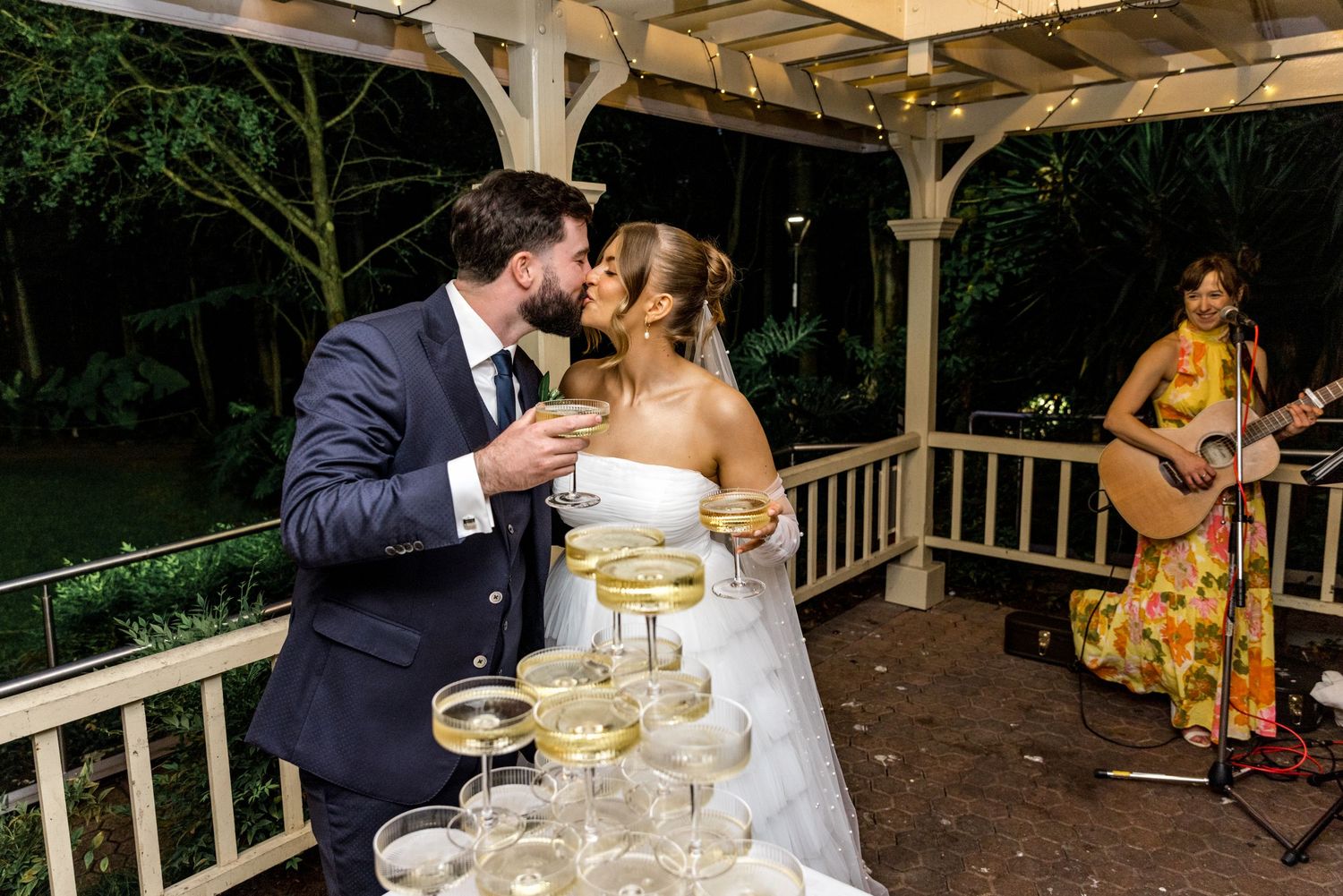 Bride and groom share a kiss behind a champagne tower at outdoor wedding reception.