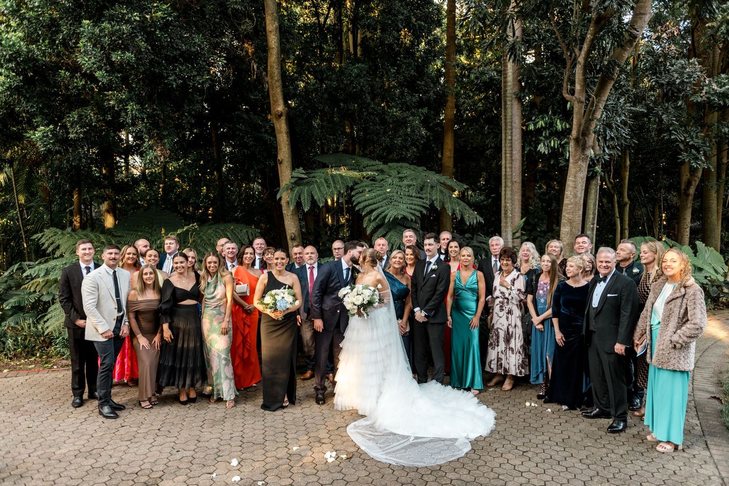 Large wedding group photo with bride and groom surrounded by family and guests in a lush garden setting.
