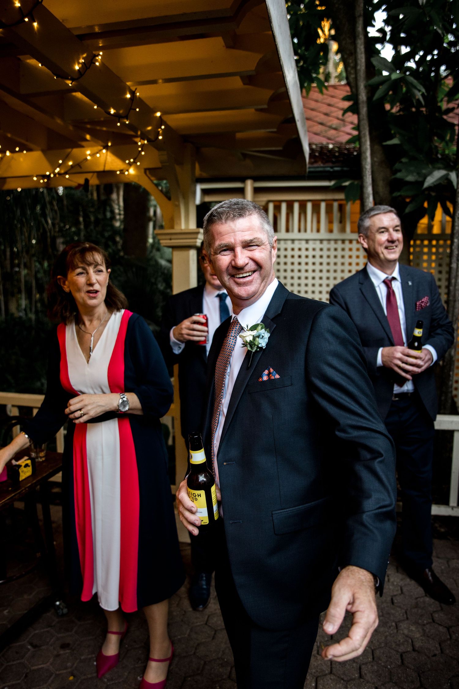 Smiling man in a suit holds a beer bottle at an outdoor wedding reception with string lights.