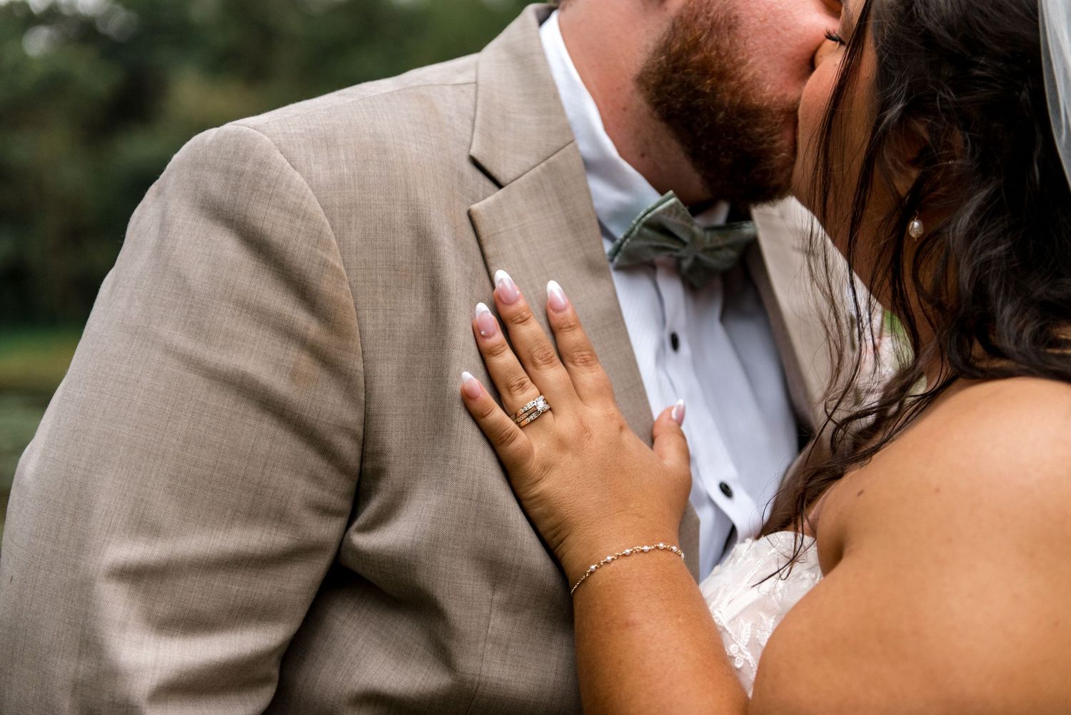 Close-up of bride resting hand on grooms tan suit jacket showing wedding ring while kissing.