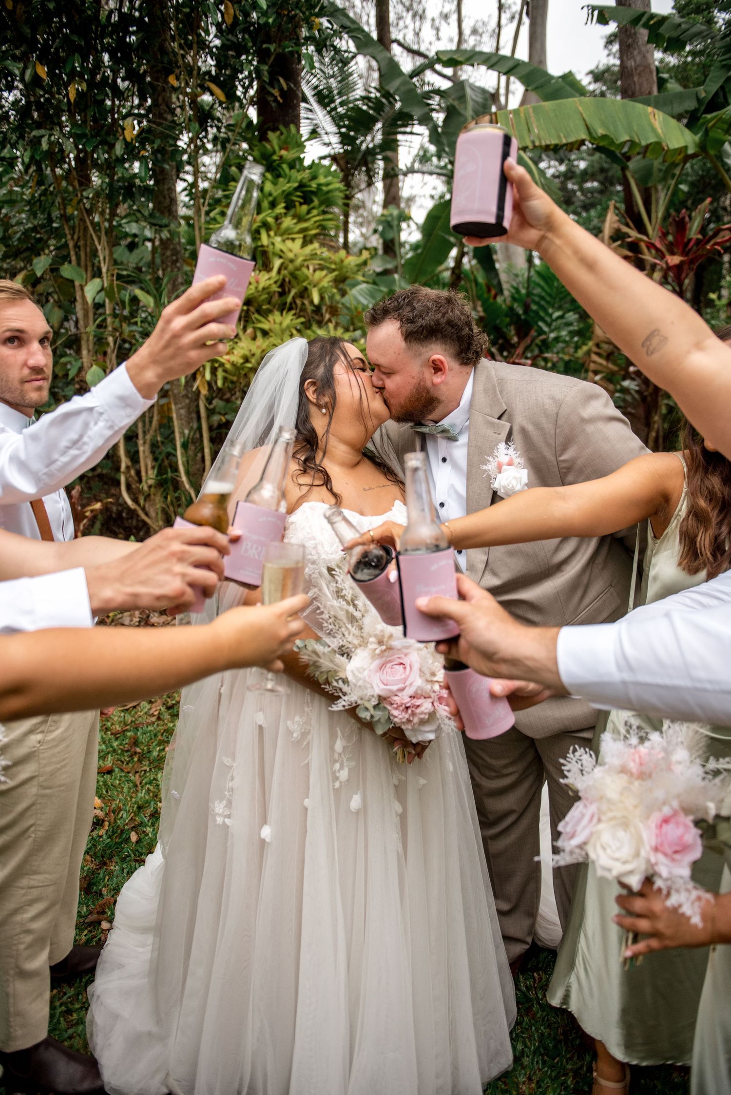 Bride and groom share a kiss while wedding party raises pink drink cans in tropical garden setting.