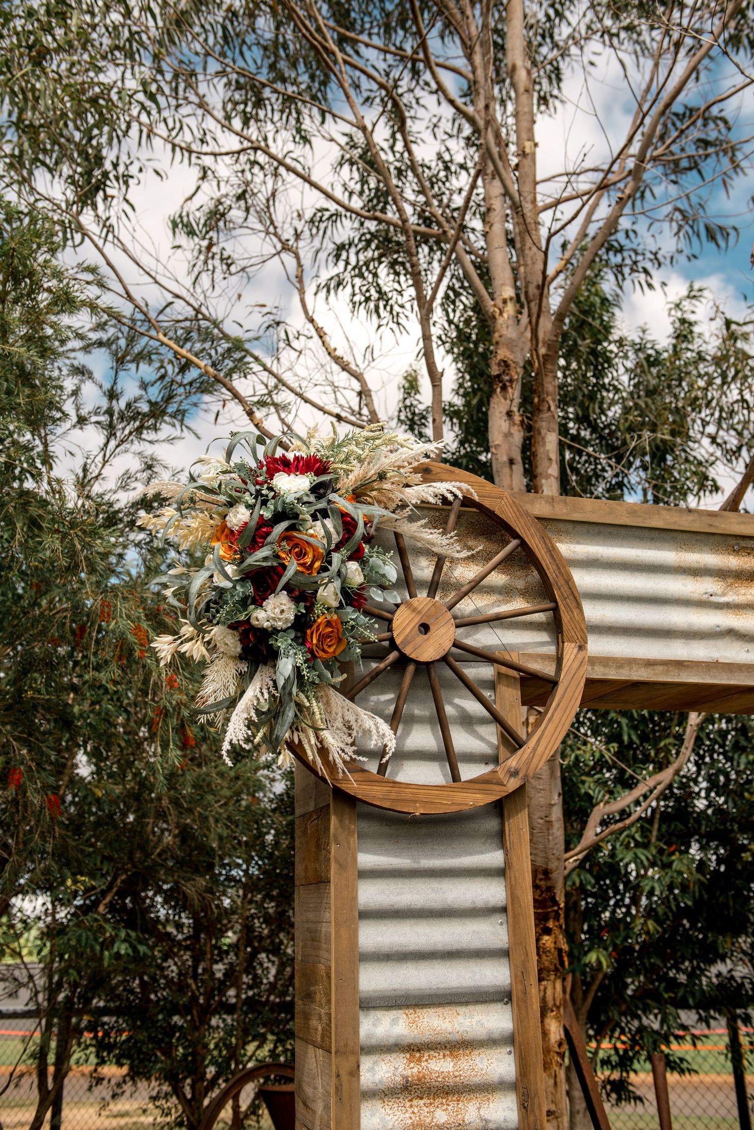 Rustic wooden wagon wheel wedding arch decorated with red, orange flowers and pampas grass against eucalyptus trees.