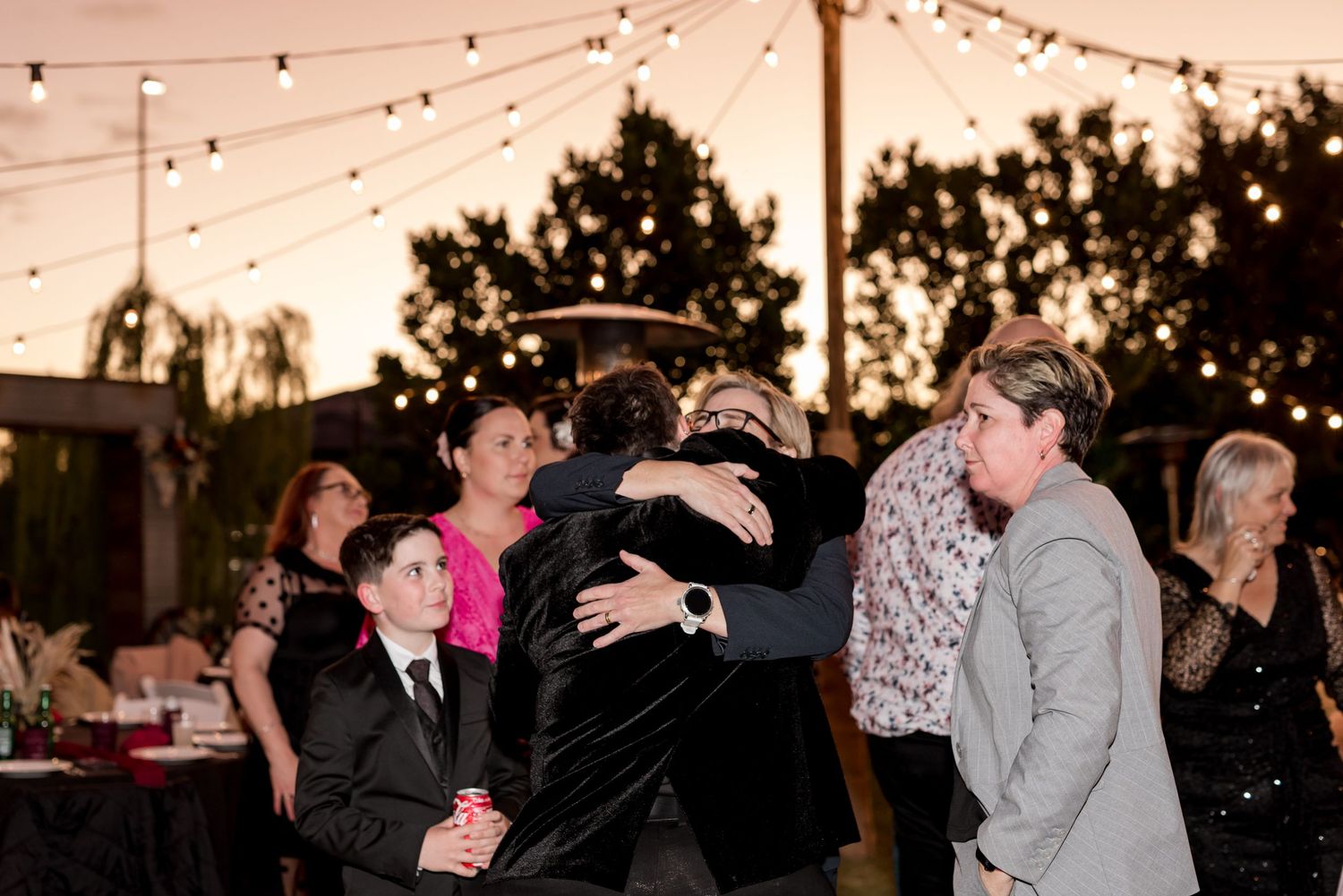 Guests share warm embraces at outdoor evening wedding reception under glowing string lights.