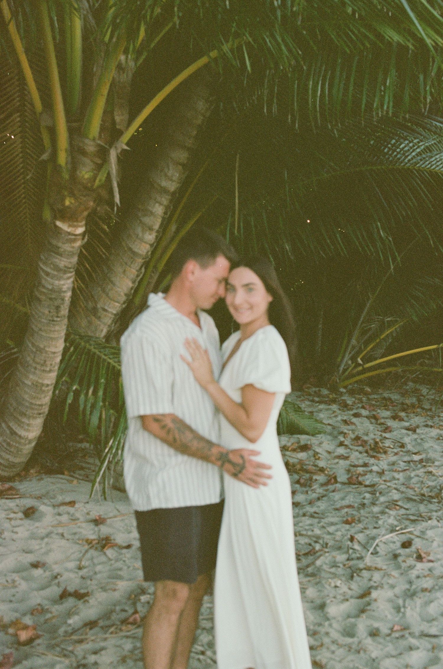 Couple embracing romantically under palm trees on sandy beach in film photography style at dusk.