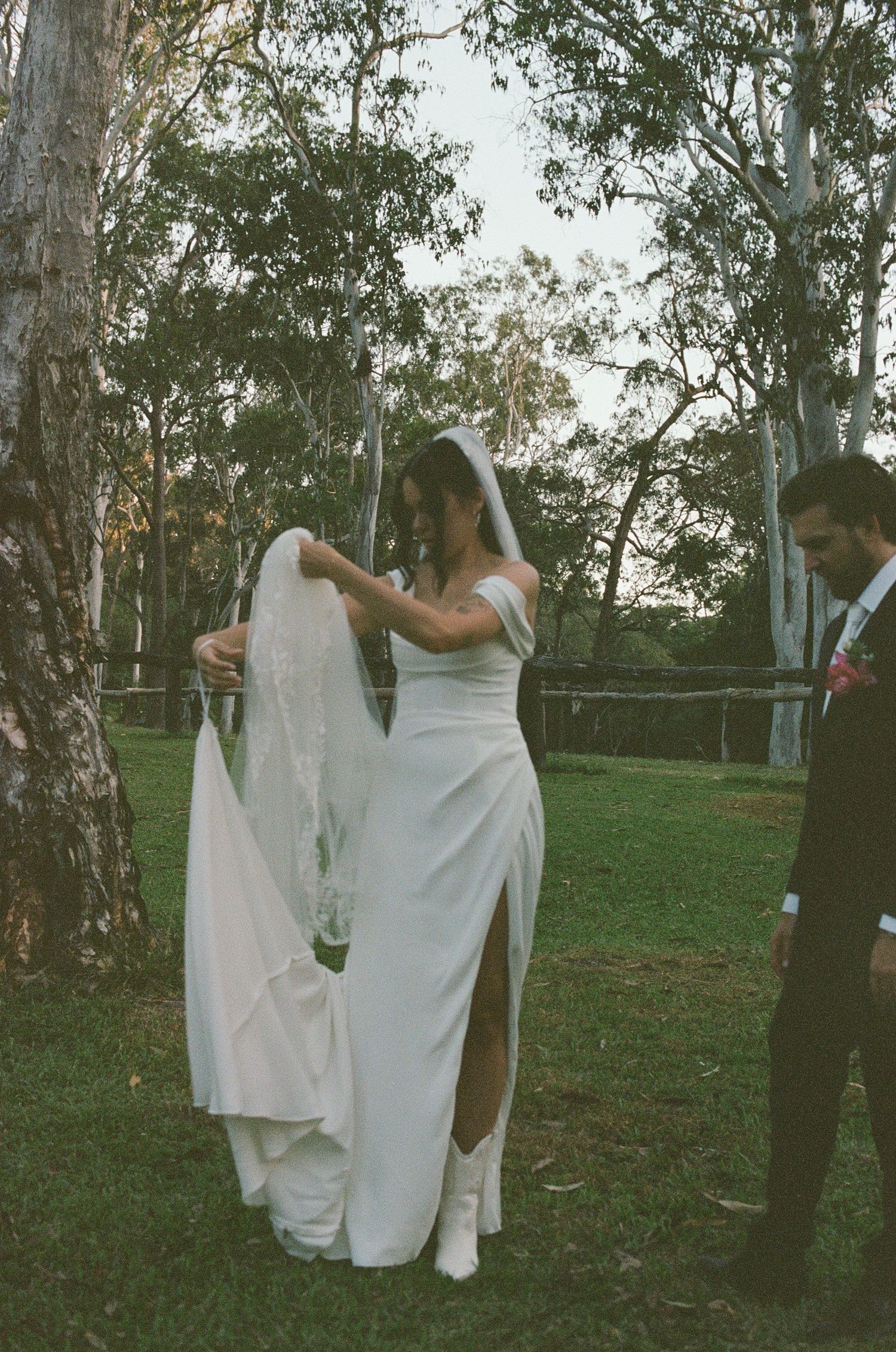 Bride removing veil in garden with groom watching near eucalyptus trees at dusk.
