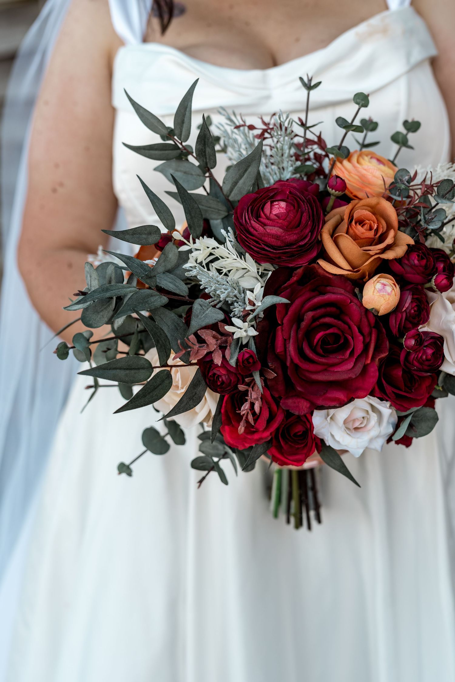 Bride in white gown holds a lush bouquet of deep red, orange, and cream roses with eucalyptus.
