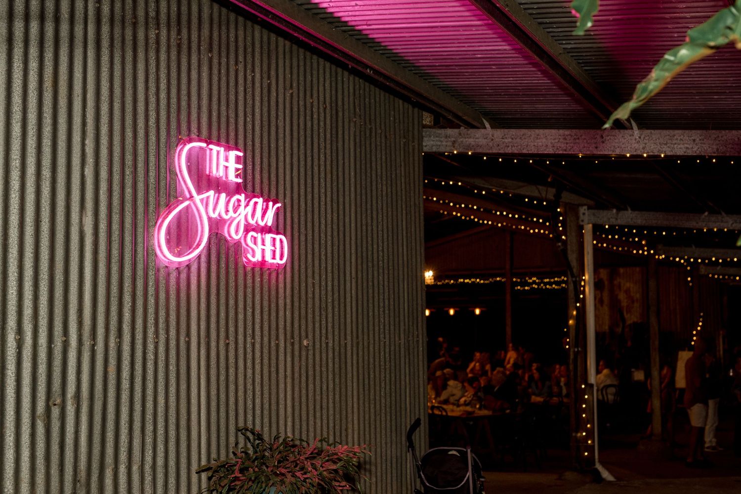 The Sugar Shed venue pink neon sign glowing at night with fairy lights on corrugated walls.