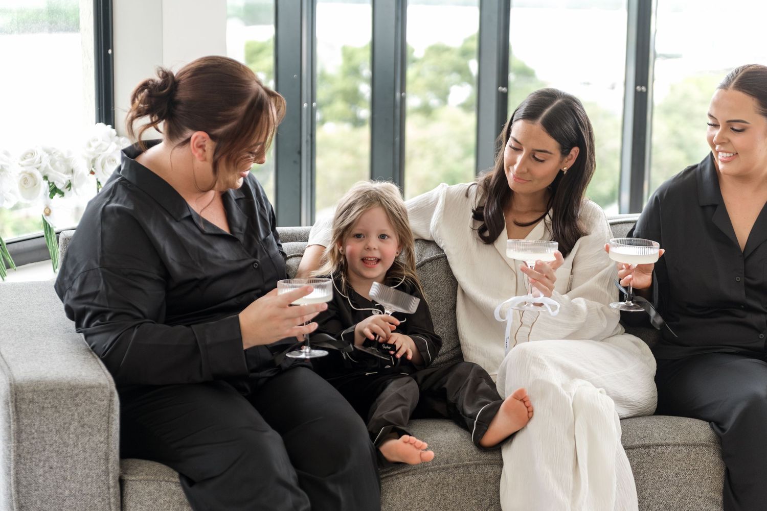 Four women and a child in satin pajamas laugh together on a grey sofa holding champagne glasses indoors.