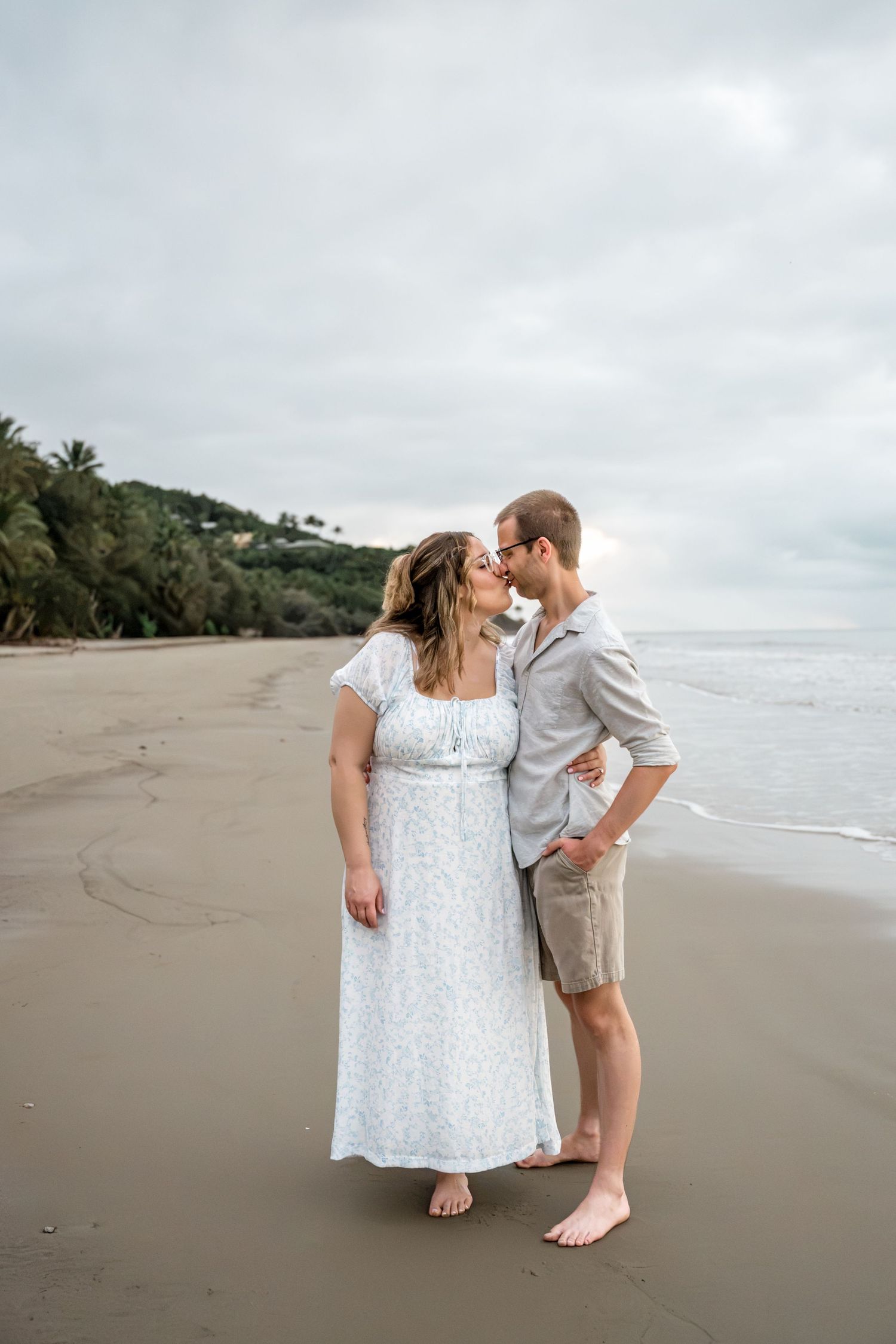 Couple sharing a kiss on a wide sandy beach with ocean waves and tropical hills behind them.