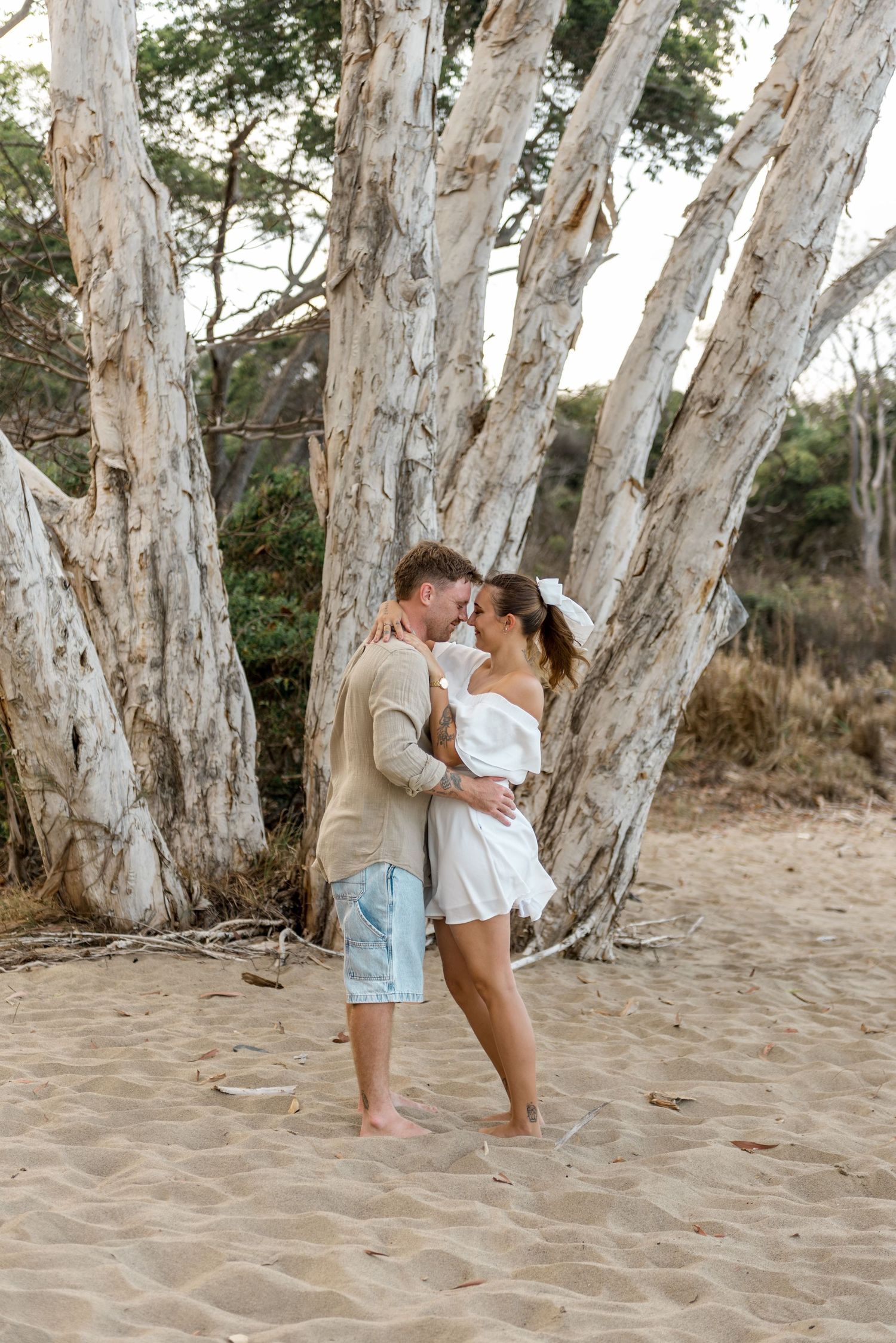 Romantic couple sharing an intimate moment barefoot on sandy beach among white paperbark trees.